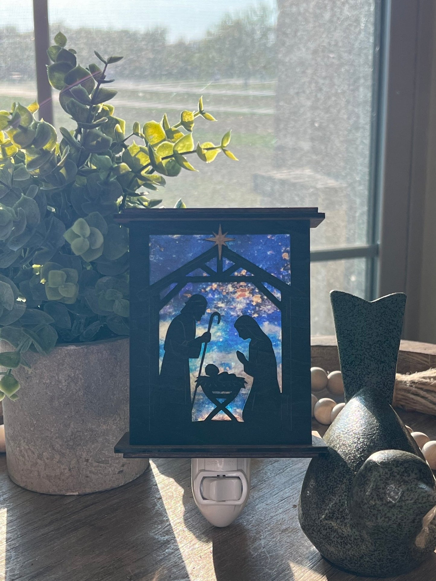 Rustic nativity nightlight lantern made of black wood and golden oak stained acrylic standingup with front view and sunlit.