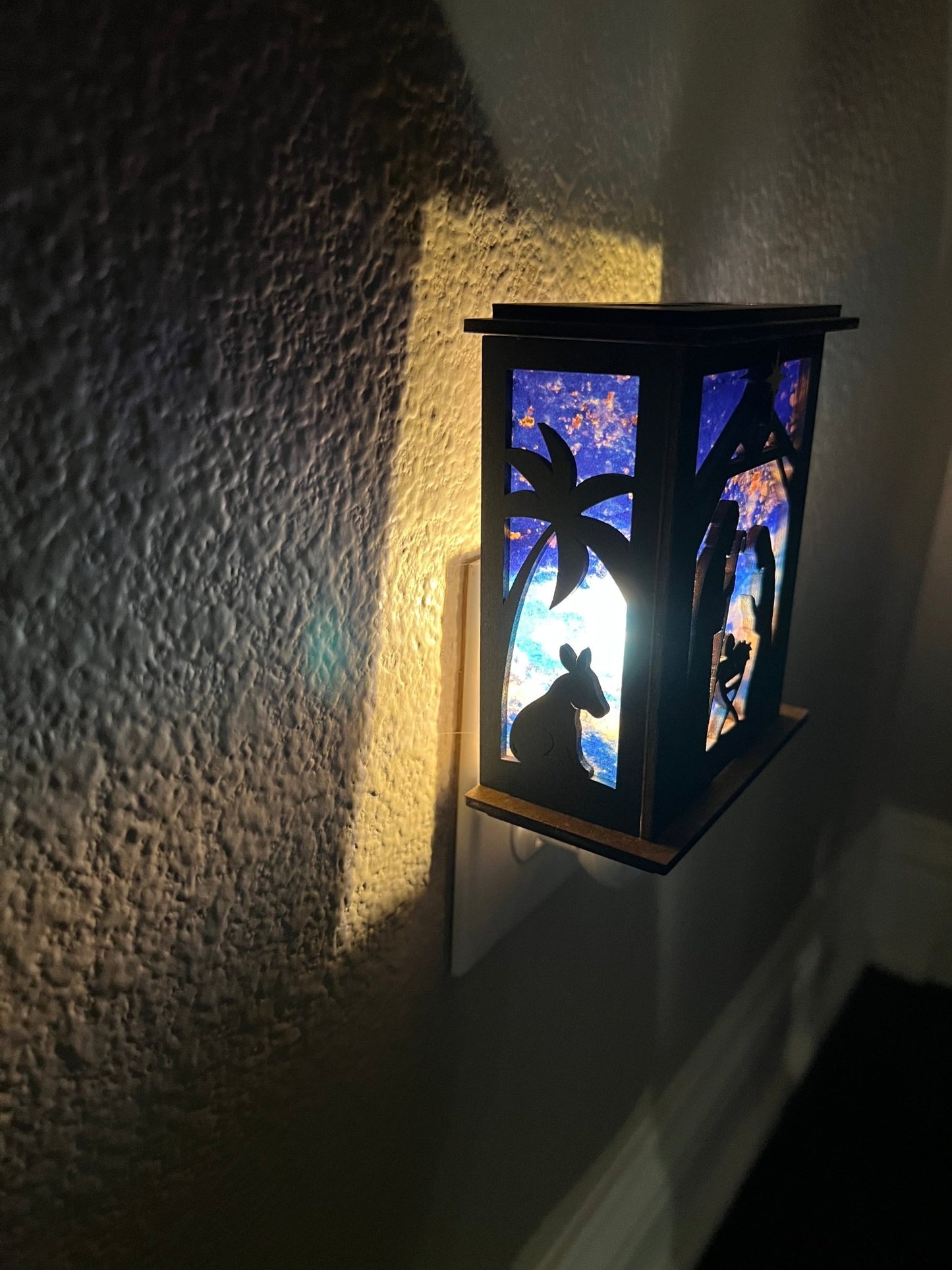 Rustic nativity nightlight lantern made of black wood and golden oak stained acrylic plugged in with left view and lit.